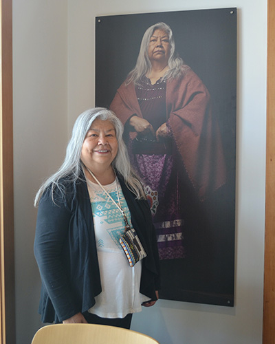 A woman with long gray hair poses in front of a portrait of herself.