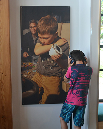 A boy wears an audio headset while standing in front of a large photo of a boy and his dad.
