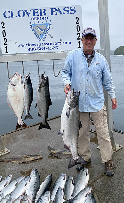 David Eaton stands on a harbor dock holding a large salmon, with several large fish on hooks behind him and about ten more on the ground in front of his feet.