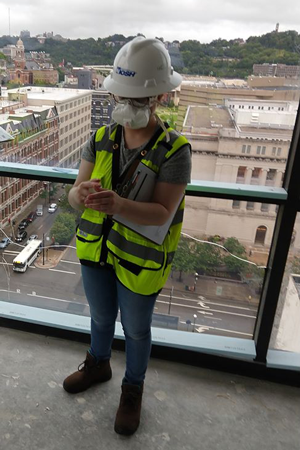 DEOHS master's student Hannah Echt wearing a hard hat, respirator, and work vest, standing inside a building in front of a window overlooking city streets