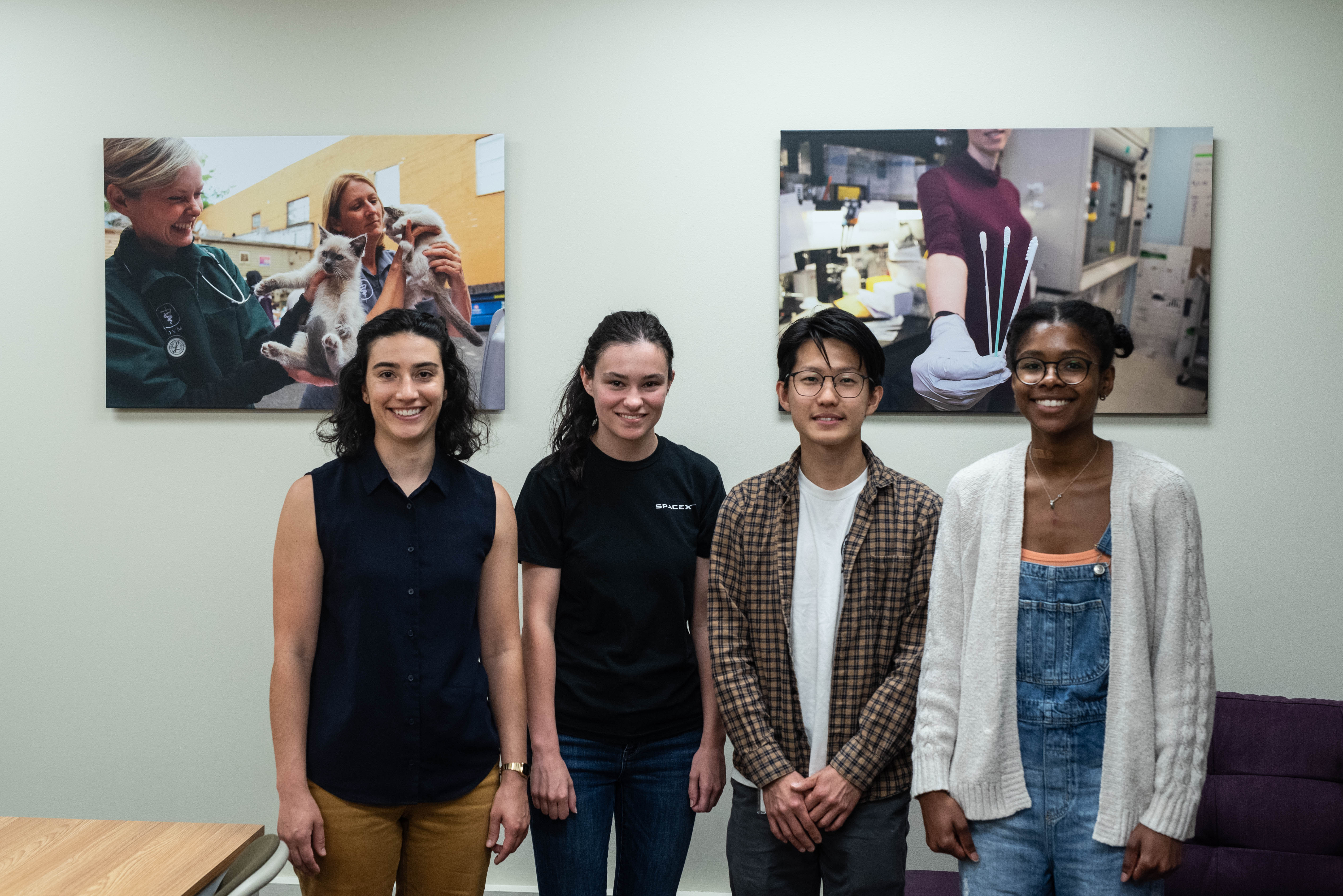 A woman with dark hair and a blue shirt stands with 3 UW students against a wall near some framed photos.