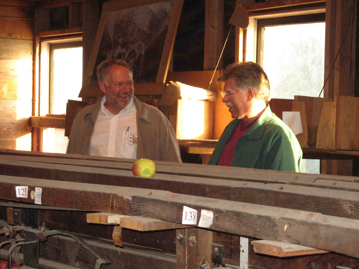 Two men talking in a barn near a pile of lumber with an apple sitting on top.