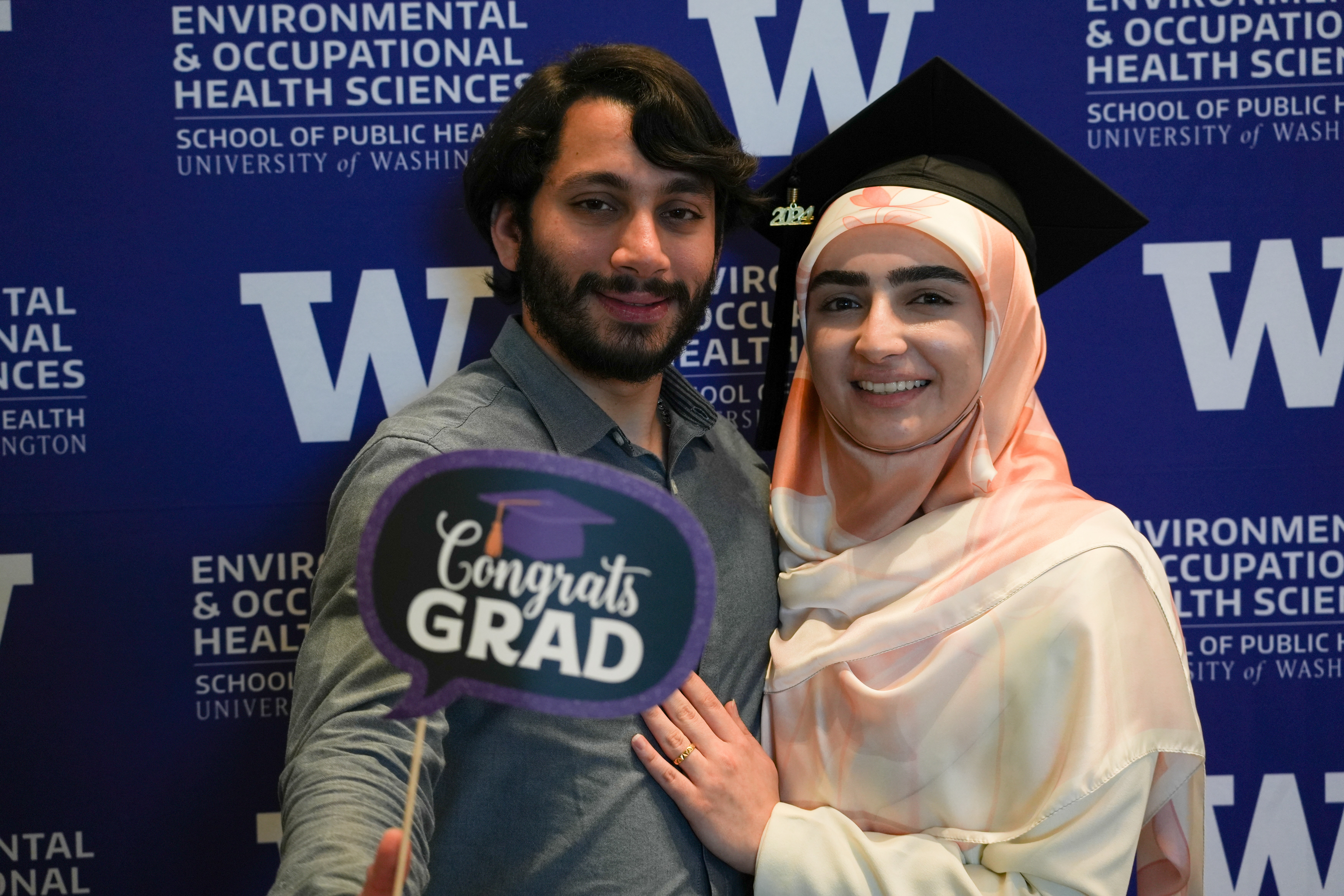 One student poses in a graduation cap and another person poses with her holding a photo booth prop that says Congrats Grad. They stand in front of a purple and white backdrop that says Environmental and Occupational Health Sciences School of Public Health University of Washington. 