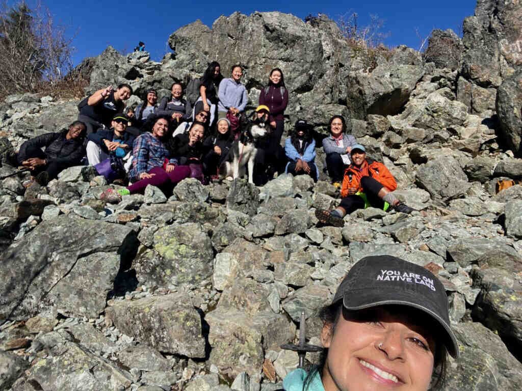 A group of about 16 hikers of color pose together on top of a mountain. In the foreground is a woman snapping a selfie wearing a shirt that says "You are on native land."