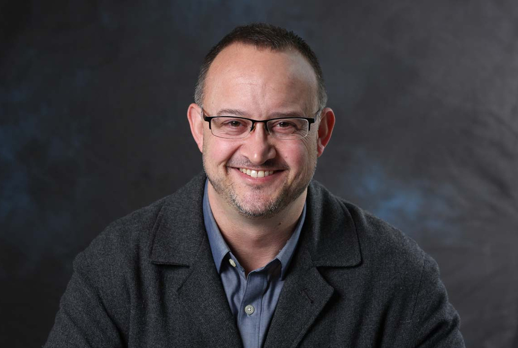 Jeremy Hess smiles at the camera. He wears a charcoal gray blazer and blue shirt as well as glasses. A grayish blue background is behind him.