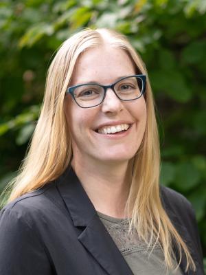 Headshot of Emily Hovis smiling and wearing glasses outside with greenery in the background.