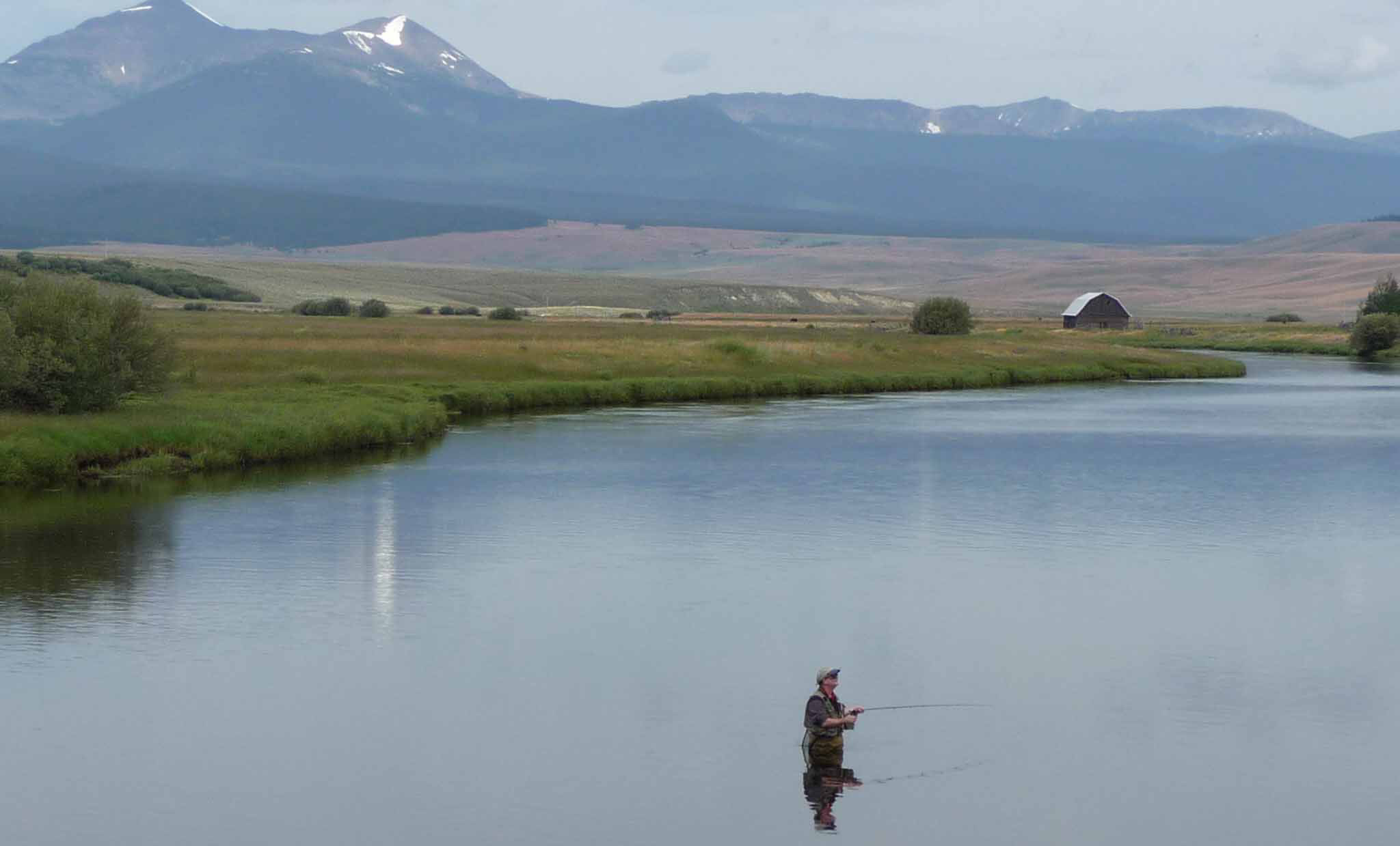A man fishes in the middle of a river surrounded by farmland and mountains.