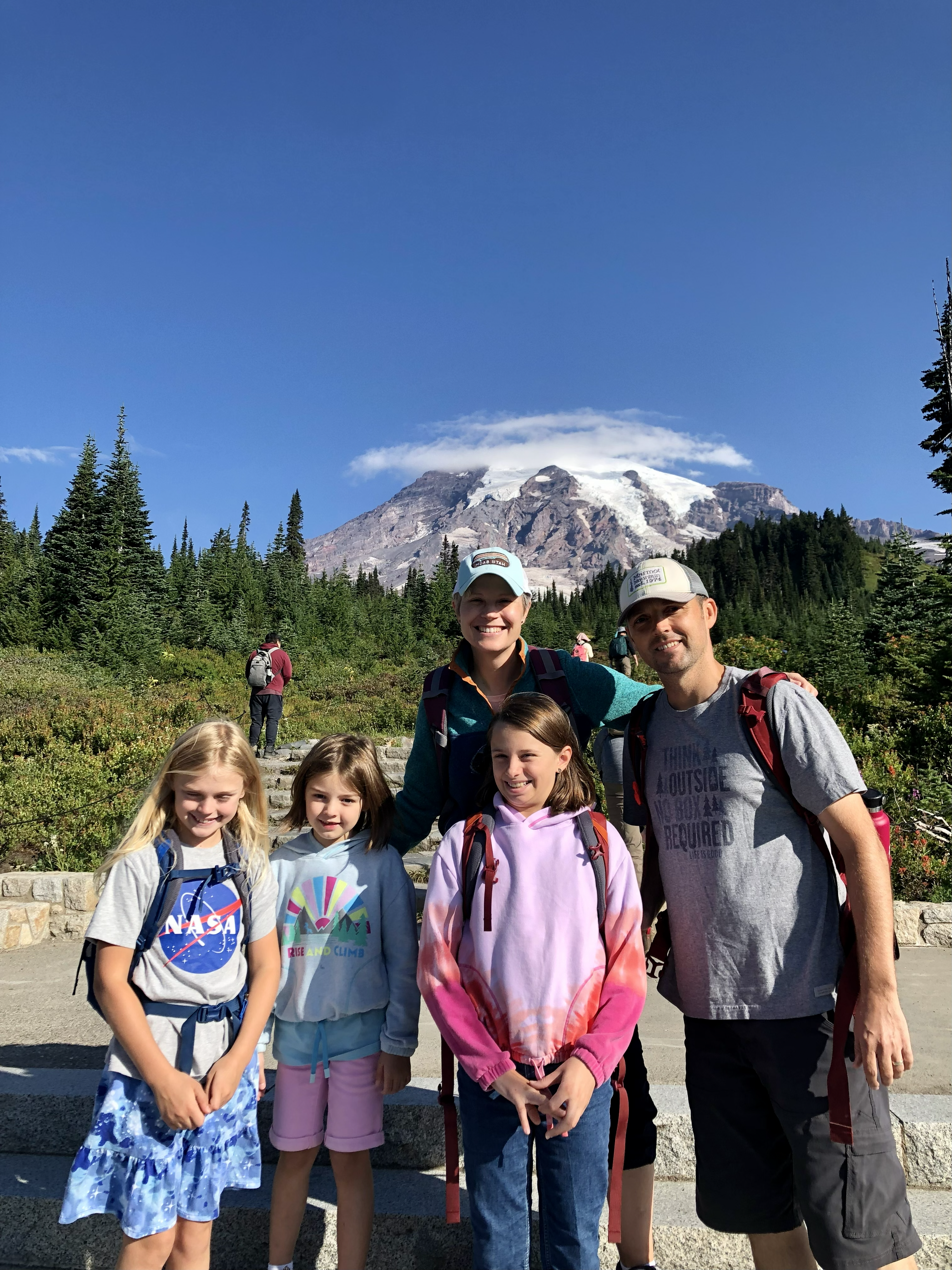 A family including a man (Mark Robinson), a woman and three children stand together smiling in front of a mountain with a snowy peak and a cloud hovering on top of the peak.
