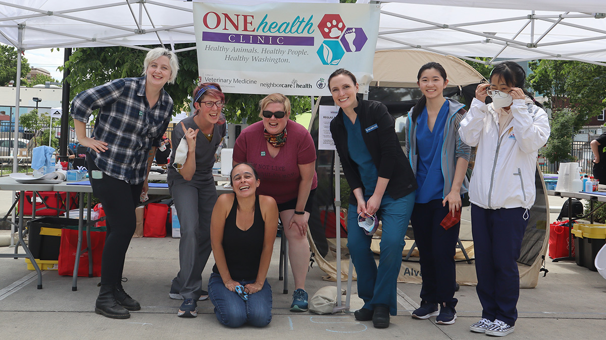 A group of people pose under a tent near a sign that reads One Health Clinic.