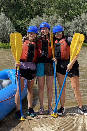 Three women stand with paddles in front of a river raft and river.