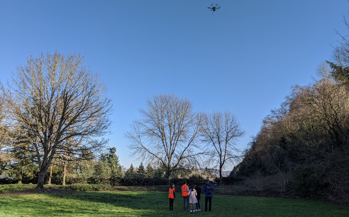 A group of five people watch a drone flying in the sky above a field and trees.
