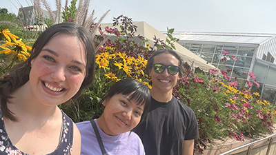 Two women and a man stand together smiling in front of a bed of flowers.