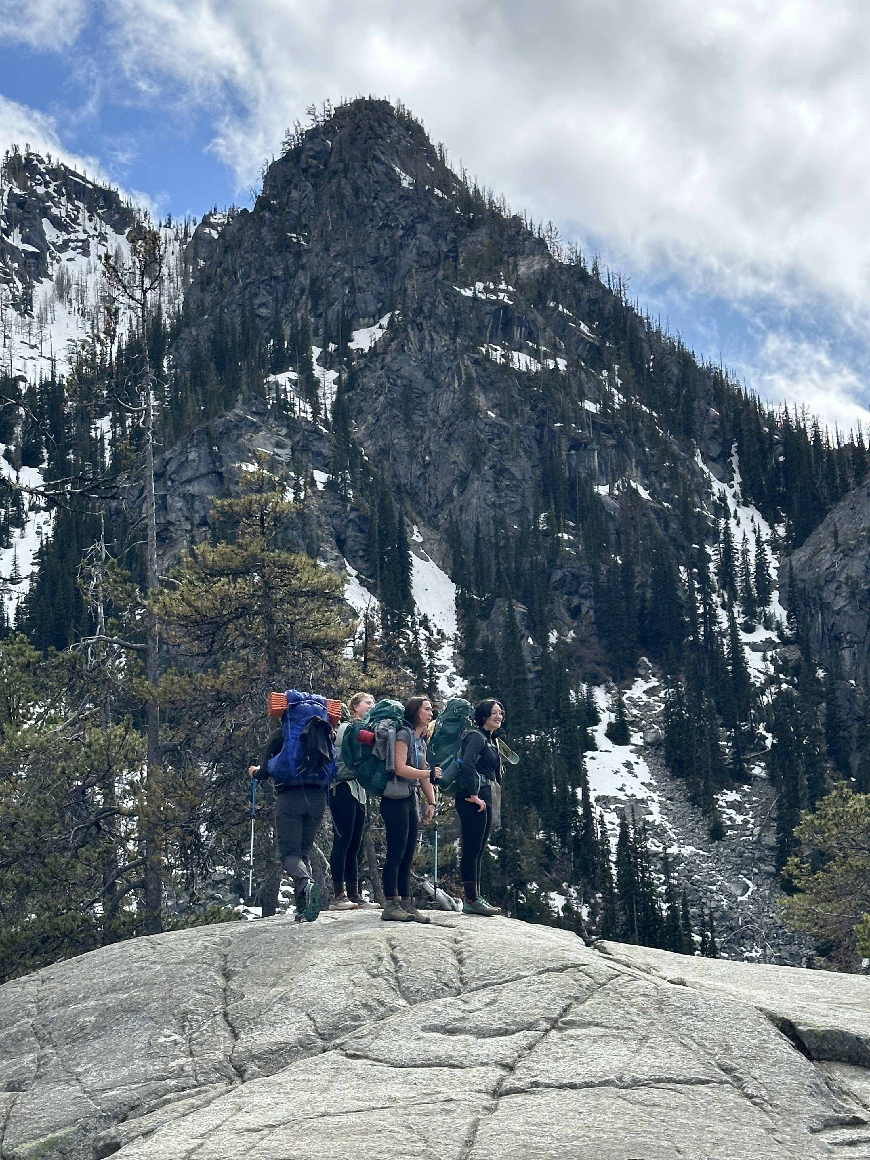 Four people with backpacking packs stand together on a rock face with mountains in the background.