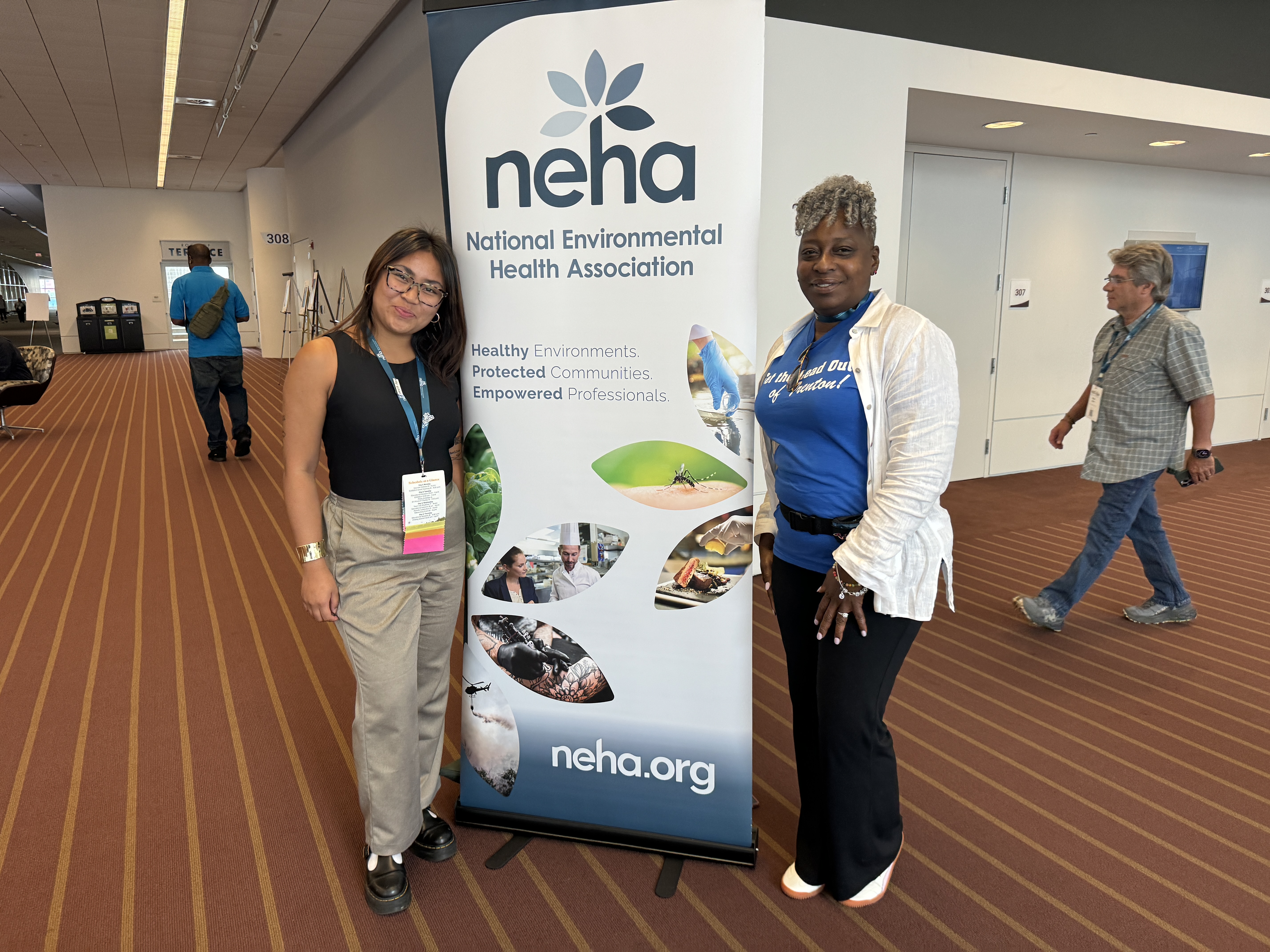 Maegan stands to the left of a pop up sign in a conference lobby. On the right is Shereyl Snider on the right side of the pop up sign. Both smile at the camera. The sign has a white and blue background with text at the top that says "neha National Environmental Health Association" text below is unreadable. Pictures in leaf-shaped frames show people in a kitchen setting, a mosquito on a leaf, a hand with a medical glove and a tattoo artist. 