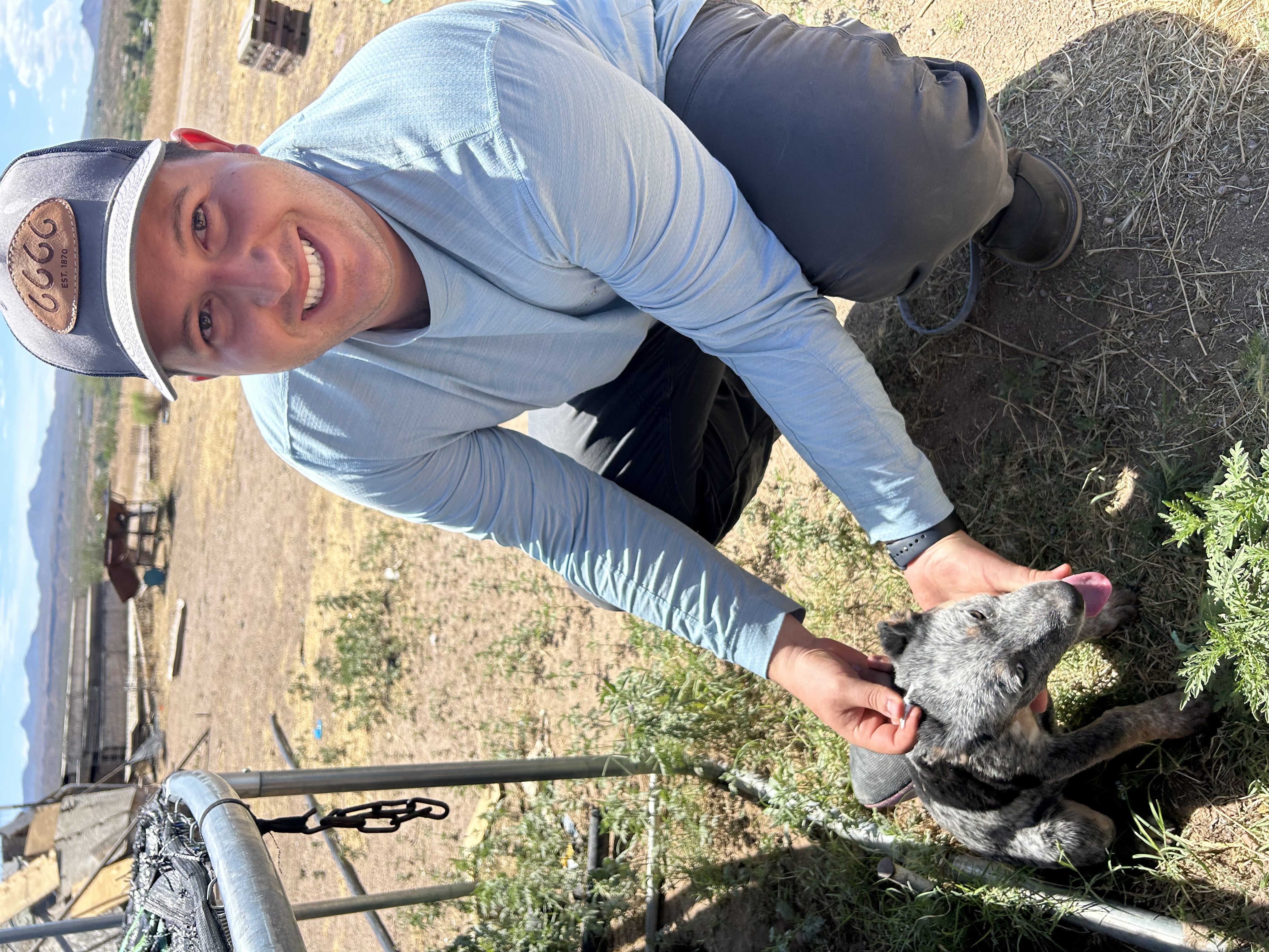 Jorge crouches next to a puppy that is gray, black and brown. Jorge and the puppy are outside and there are hills and blue sky in the background. Jorge wears a blue long sleeve shirt and baseball hat while smiling at the camera.