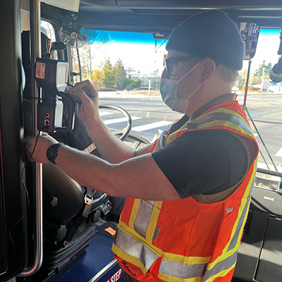 A man wearing a cap and an orange vest checks air ventilation near the driver's cab inside a bus.