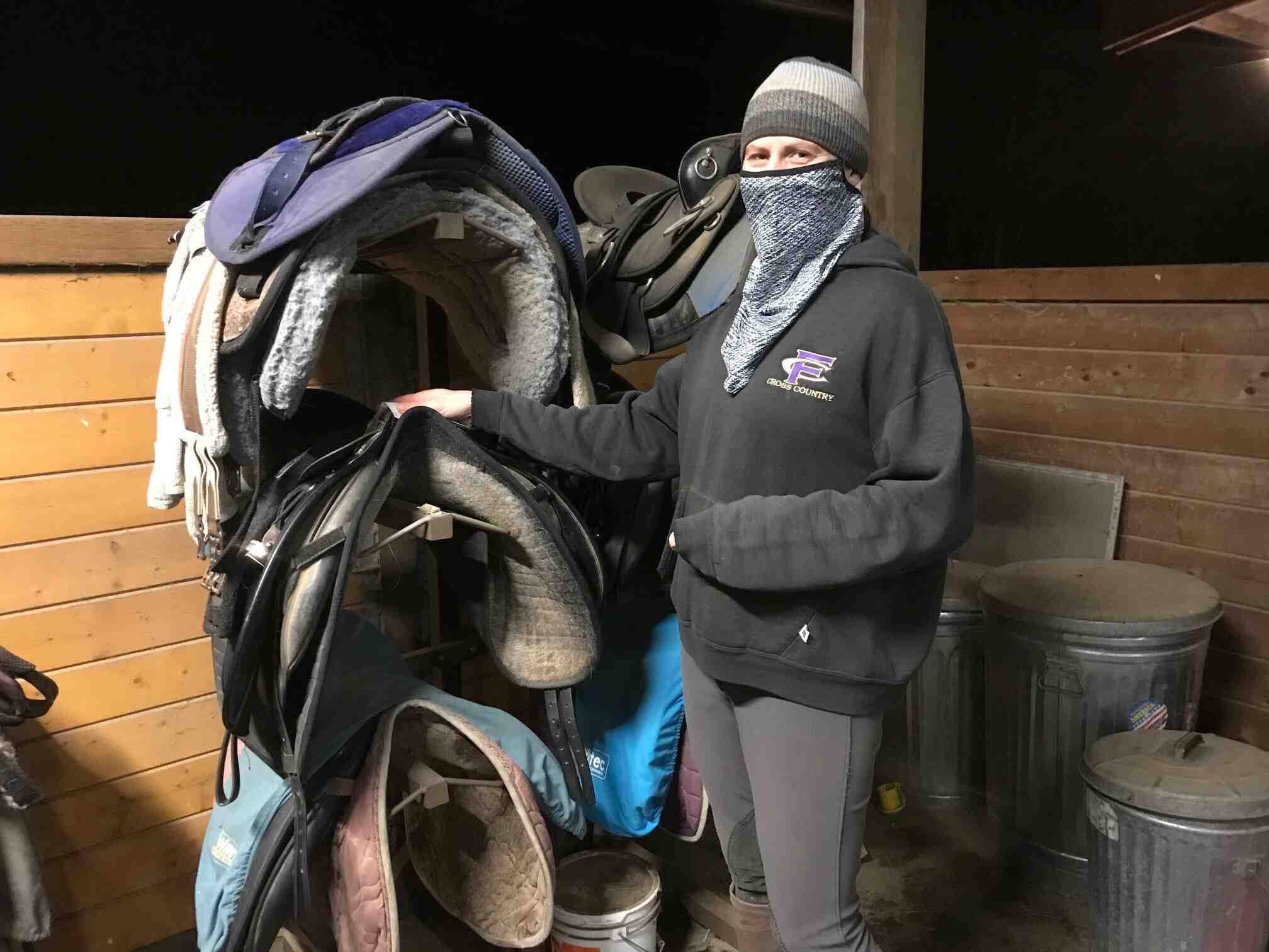 A woman with a mask stands next to saddles in a horse stall.