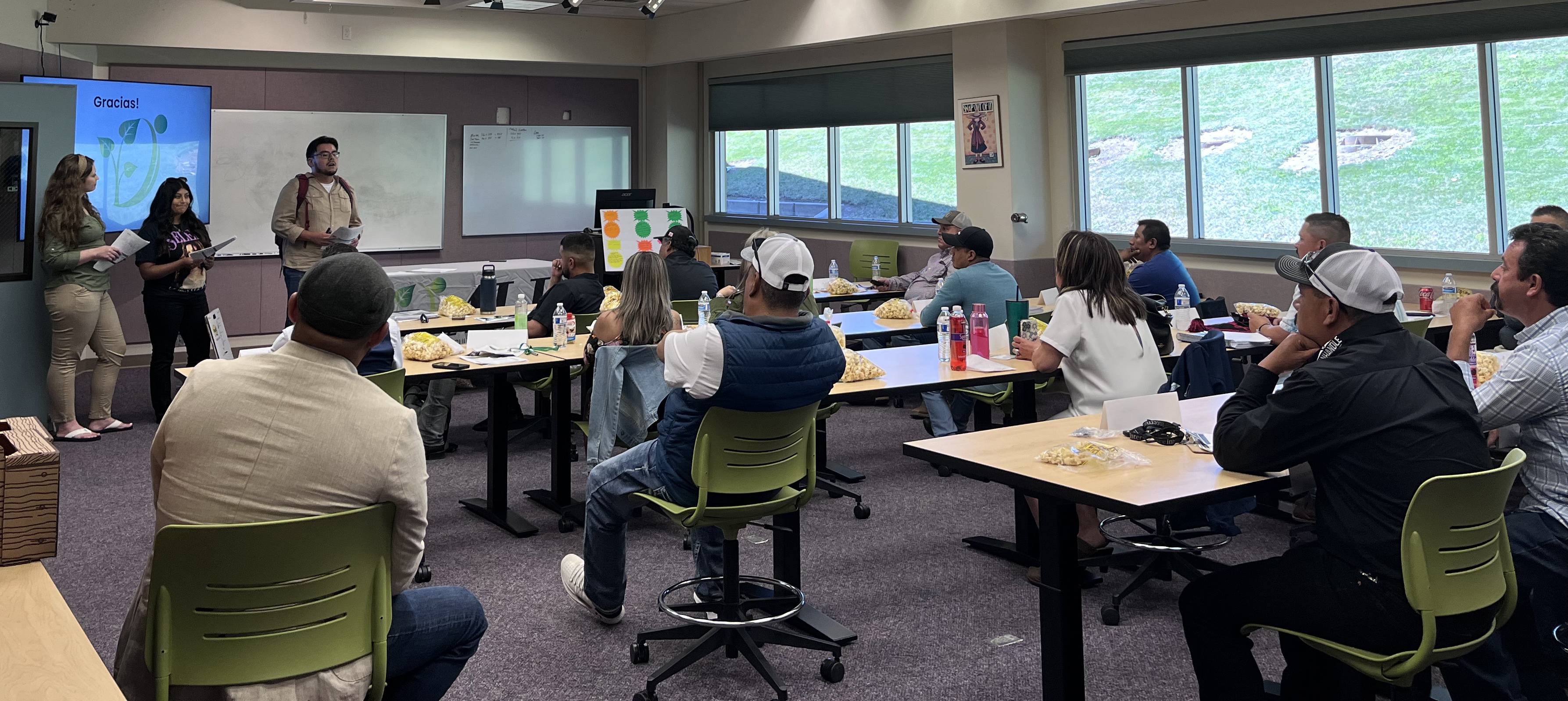 A group of people sitting in chairs watch a man talking to them from a podium by a whiteboard.