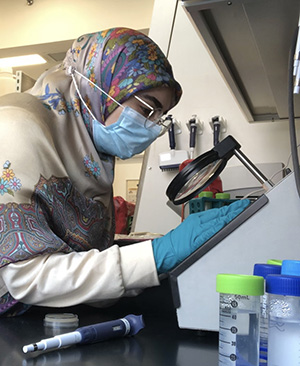 A woman in a hijab leans over a magnifying lens in a lab.