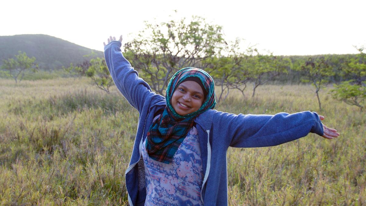 Woman in a hijab stands in an open field with trees in the background holding her arms out and smiling.