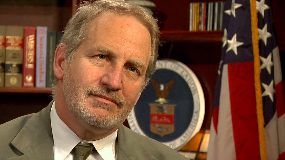 Jordan Barab sits in front of a book shelf with the American flag on the right side of the picture. He wears a tan suit and dark tie with white shirt. He has a neutral smile and looks off to the right side of the camera.