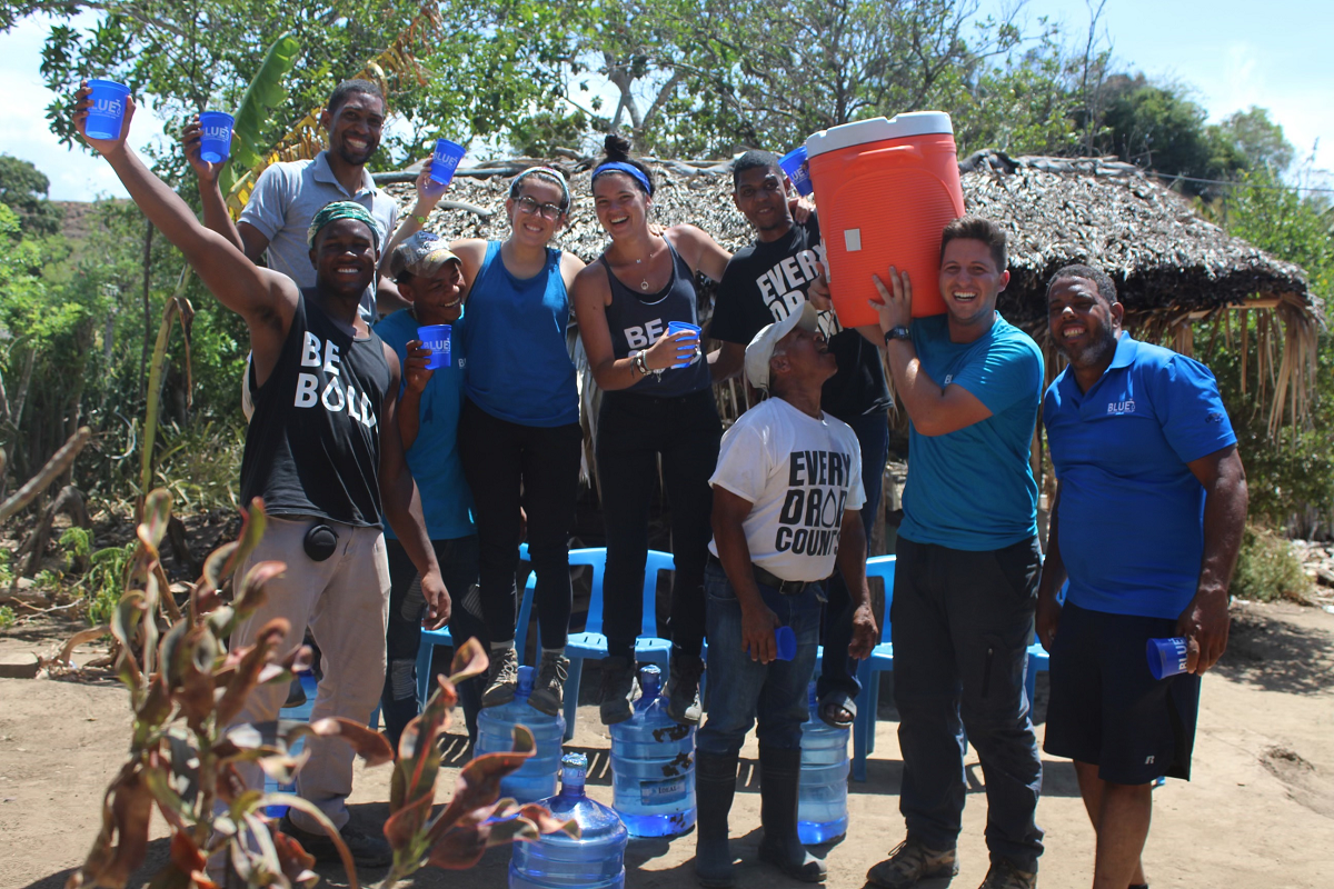 A group picture of 8 people holding up glasses of water, with t-shirts with messages including "Be Bold" and "Every Drop Counts"