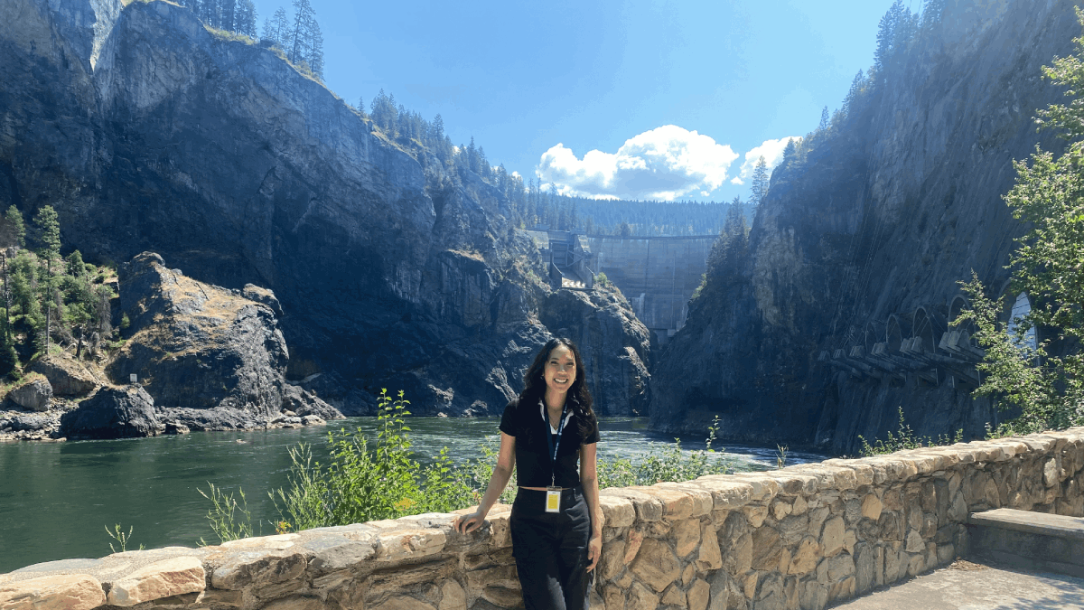 Kai stands against a stone wall in front of a dam.