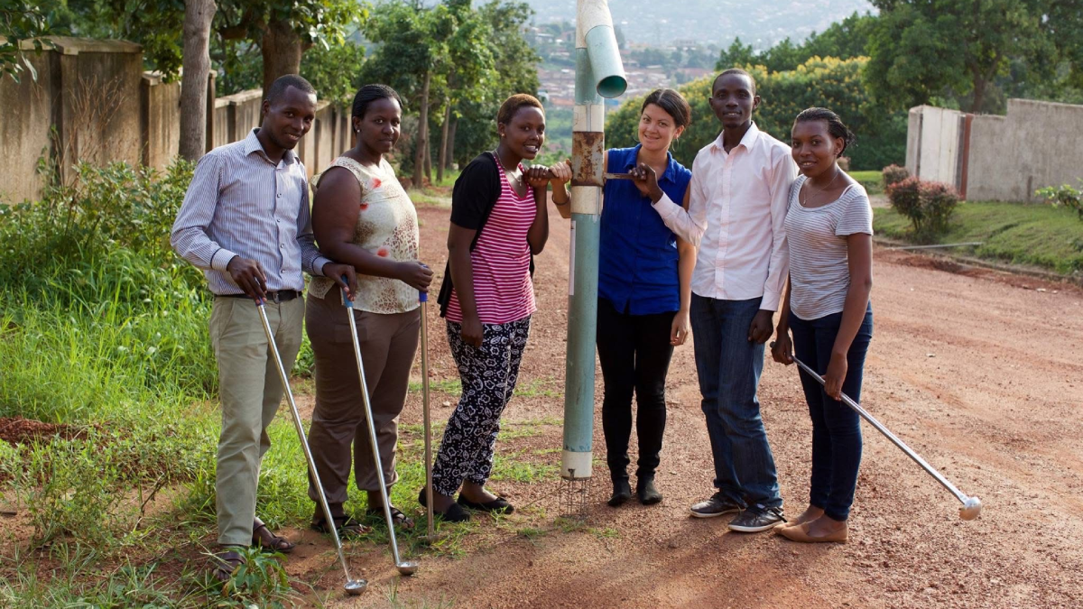 A group of six people including Sklar stand on a dirt road. In the middle of the group, Sklar and one other person hold up a PVC pipe. Several people in the group are holding golf clubs.