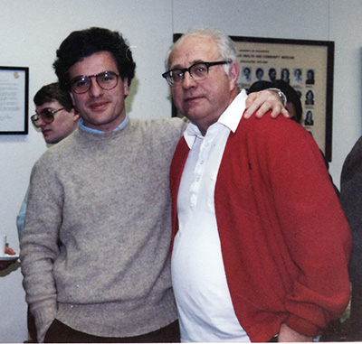 A young man in glasses stands with his arm around the shoulders of an older man in a red sweater.