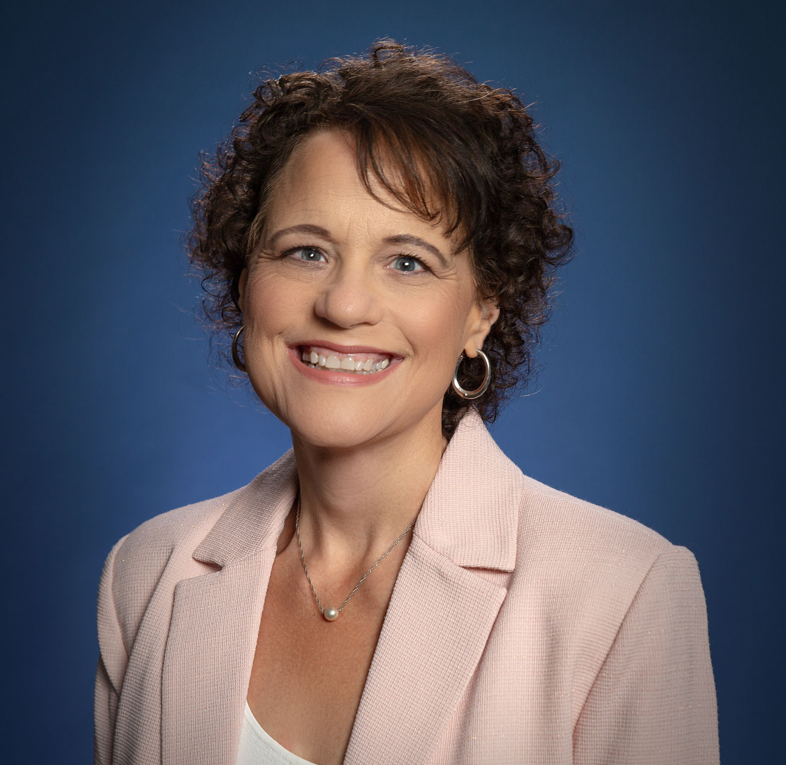 White lady with curly brown hair in a pink suit jacket with a big smile on her face. 