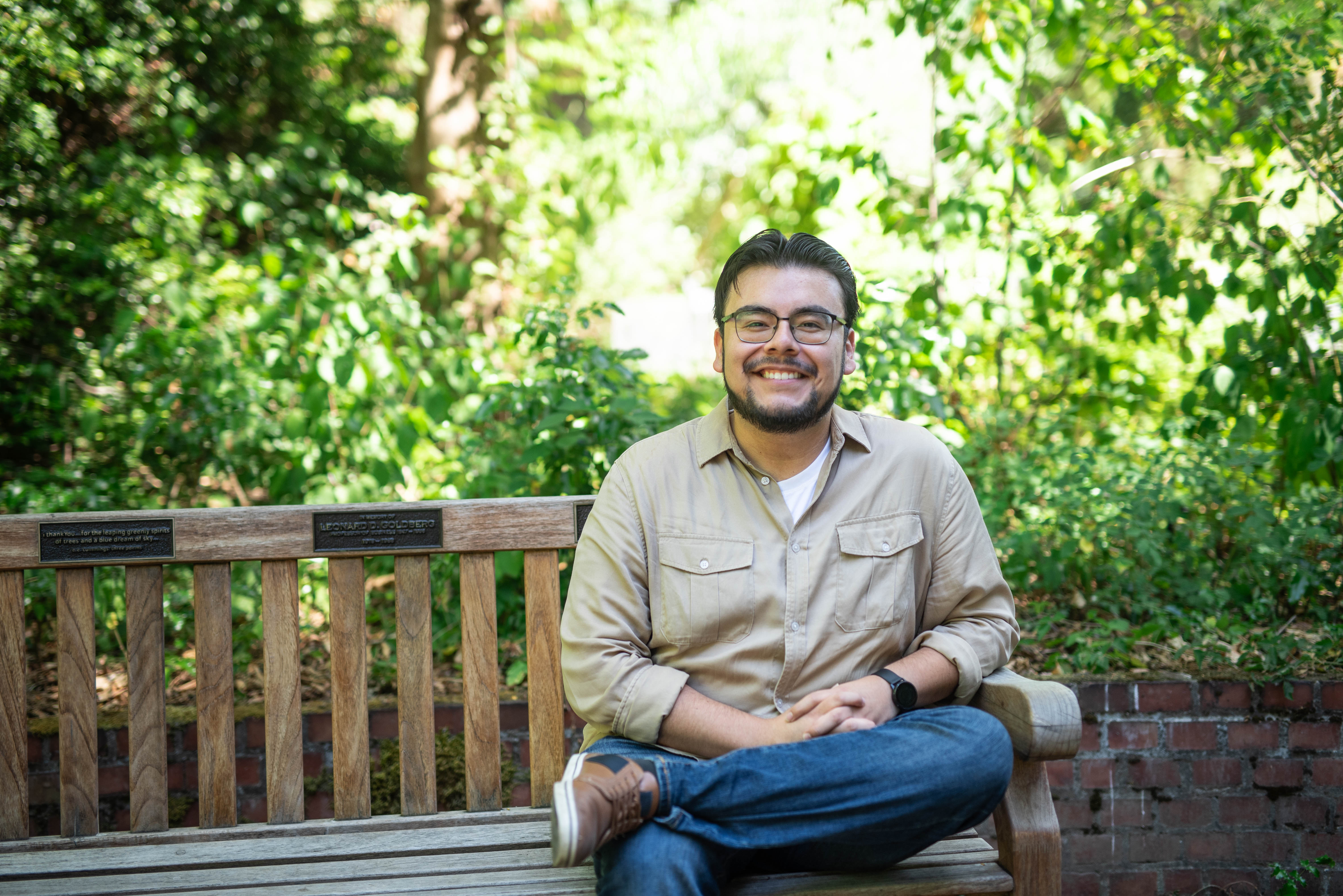 A man in glasses and a tan shirt sits on an outdoor bench.