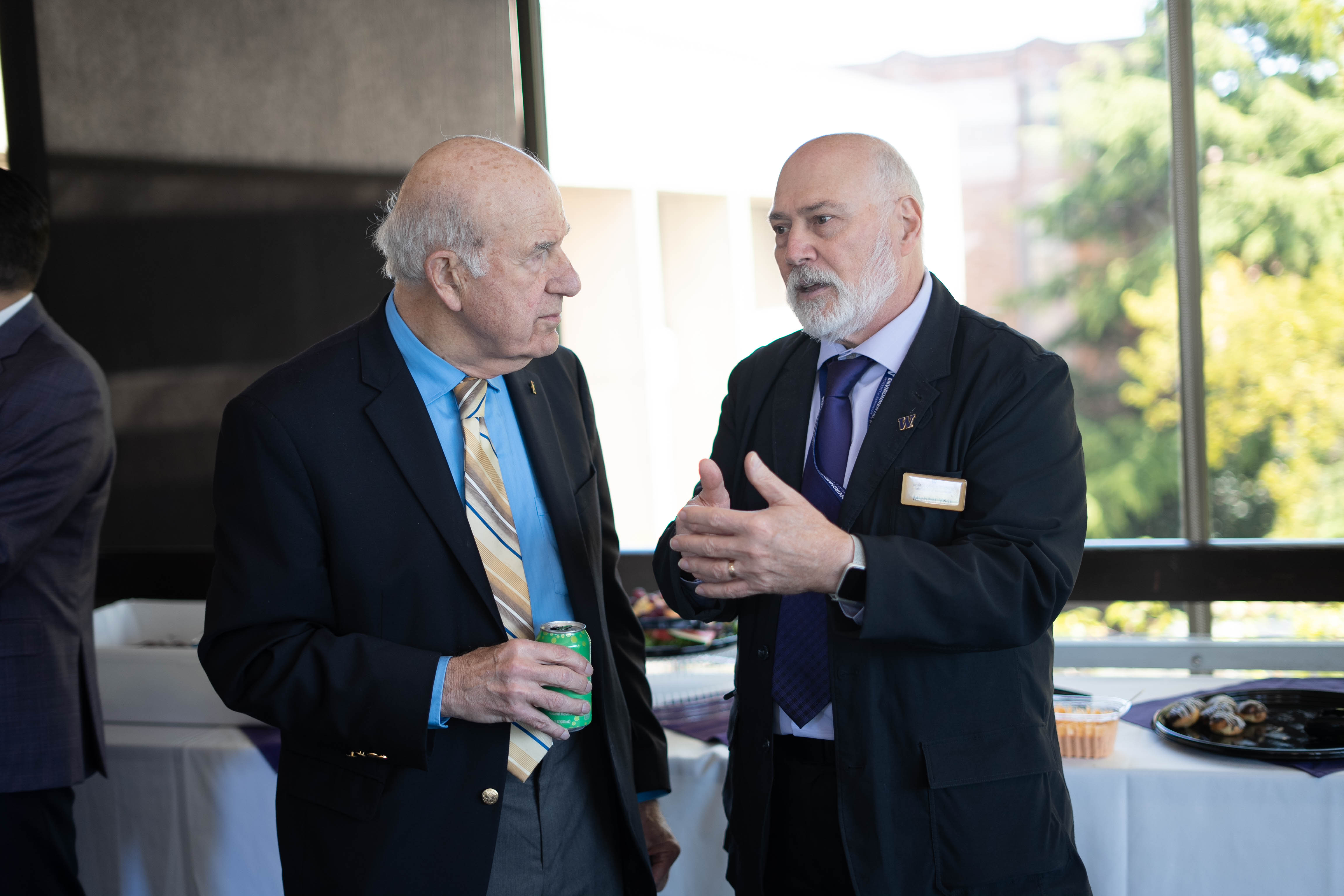 Michael Yost stands with Gil Omenn, in conversation, at a reception. Both are dressed in jackets and ties with banquet tables behind them.