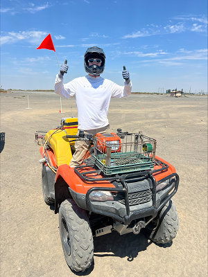 Eribac, wearing a protective helmet and goggles, stands in an all-terrain vehicle in a dusty field holding both thumbs up.