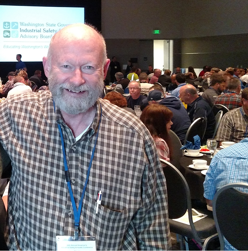 Rick Gleason stands in a crowded banquet room, smiling. He wears a button up shirt and has a lanyard around his neck with a pen in his shirt pocket.