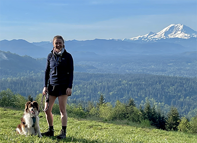 Schollaert stands with her dog on a mountain with Mount Rainier in the distance.