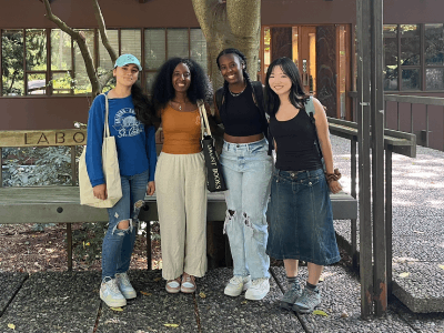 Four people stand together smiling in front of a building on campus.