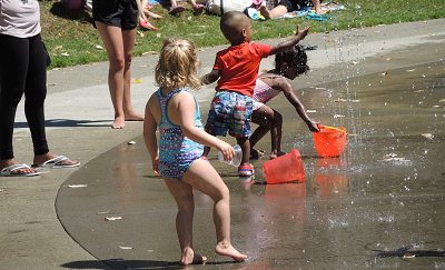 Three kids play in a spray park with buckets while two adults (with only legs visible) stand nearby