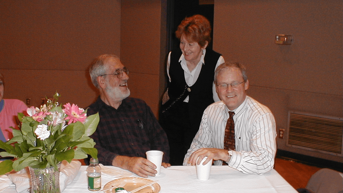 Sharon Morris smiles while leaning down to talk to two men seated at a table with paper cups and a vase of flowers.