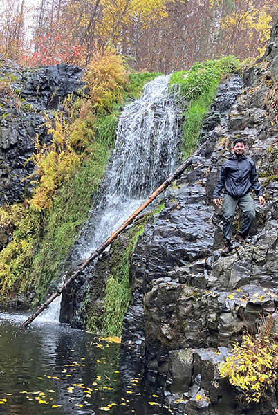 Photo of Yoni Rodriguez standing on a rock outcrop beside a waterfall and lake.