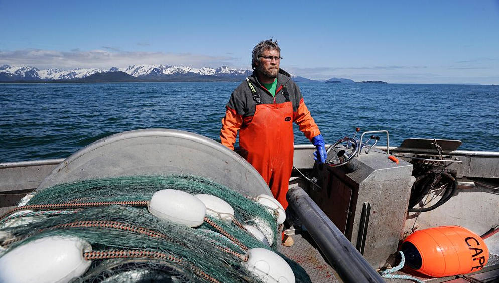 Fisherman stands on the deck of a silver, metal boat, mountains and the ocean behind him and fishing equipment around him.