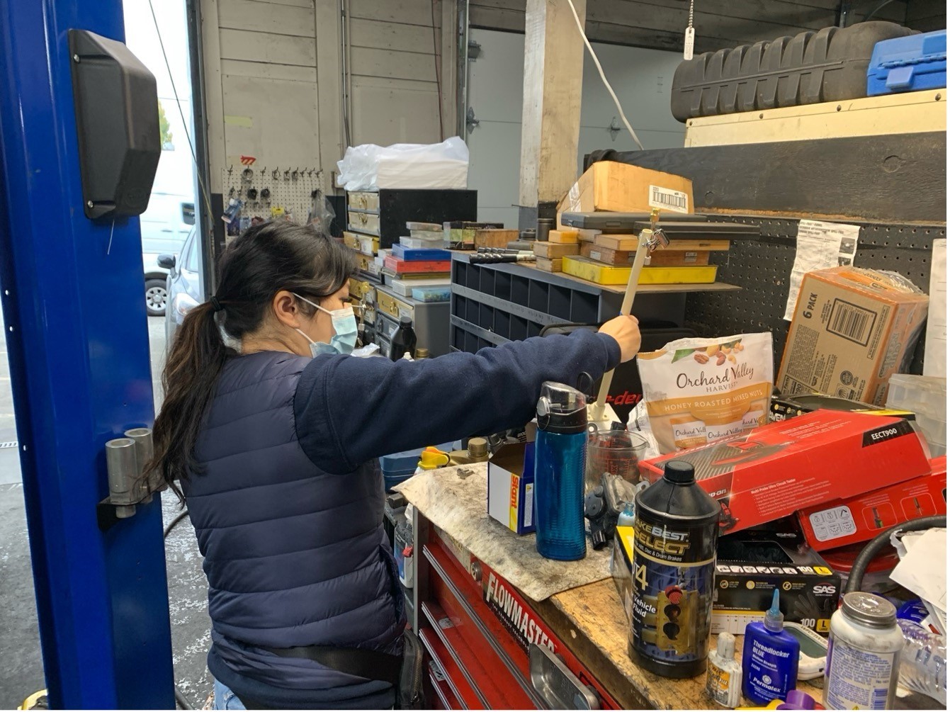 A worker stands at a work bench inside an auto repair shop. The bench has many supplies including boxes, cans and bottles, with lables that are unclear. The worker wears a face mask. In the background, the garage door is open and cars are parked in the parking lot.