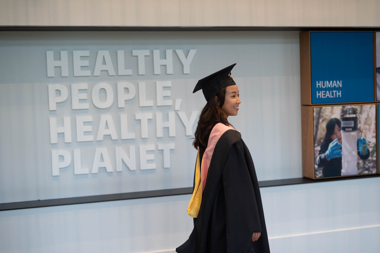 A woman in cap and gown poses in front of a sign that reads Healthy People, Healthy Planet.