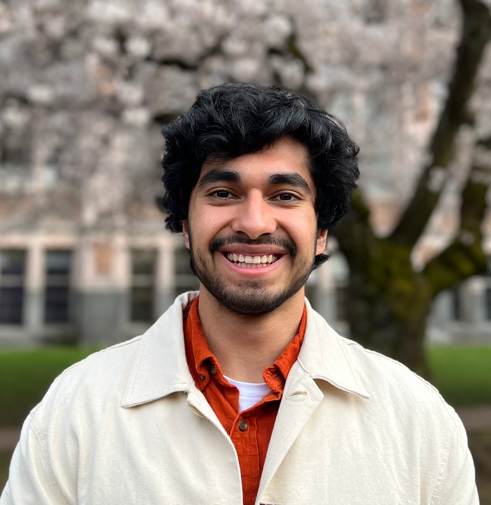 A young man with dark hair, brown eyes and facial hair, smiling with cherry blossom trees in the background. He is wearing a creme jacket and orange button up underneath.