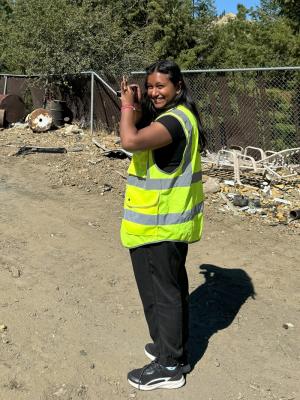 Student with long black hair and a yellow safety vest uses a camera to take pictures. Twisted metal and debris are in the background.