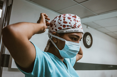 A healthcare worker wearing scrubs, a face mask and a head wrap holds arms up to their head, with a clock behind them.