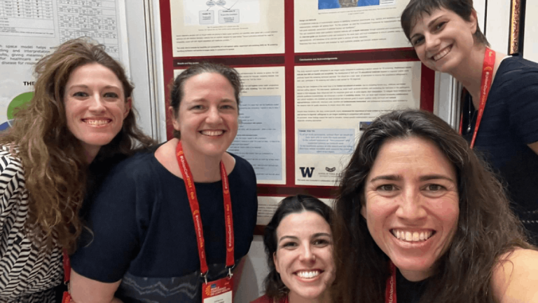 Five women stand together smiling in front of a research poster.