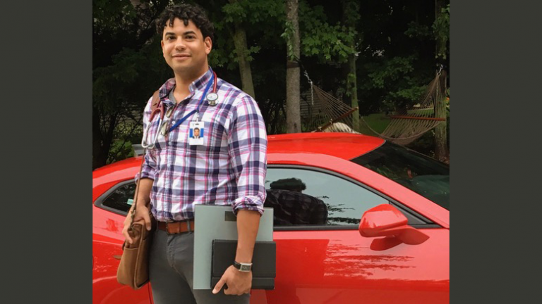 Kenleigh stands, with a briefcase on his shoulder and a stethoscope around his neck, in front of a red car.