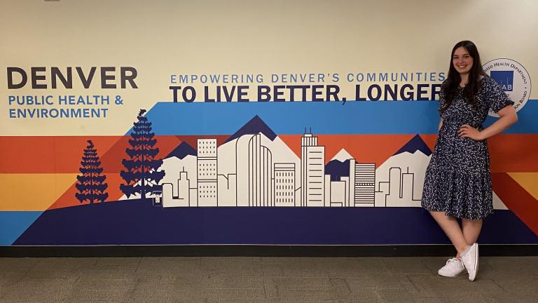 Belen Salguero stands in front of a wall mural showing Denver skyline with text: "Denver Public Health & Environment. Empowering Denver's Communities to Live Better, Longer"