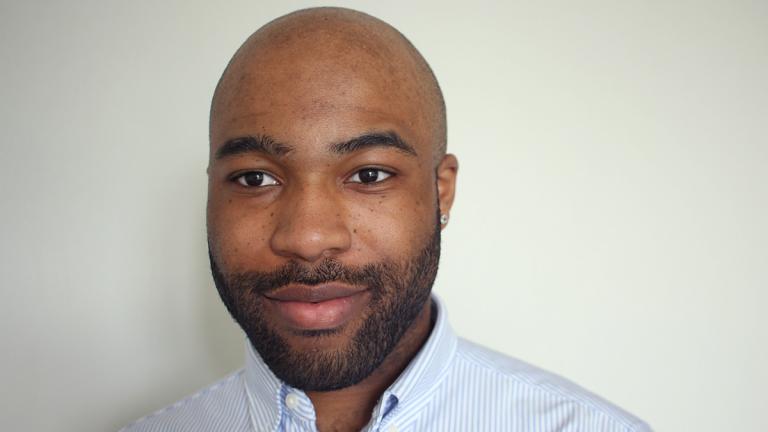 A man in a blue striped shirt poses against a white wall.