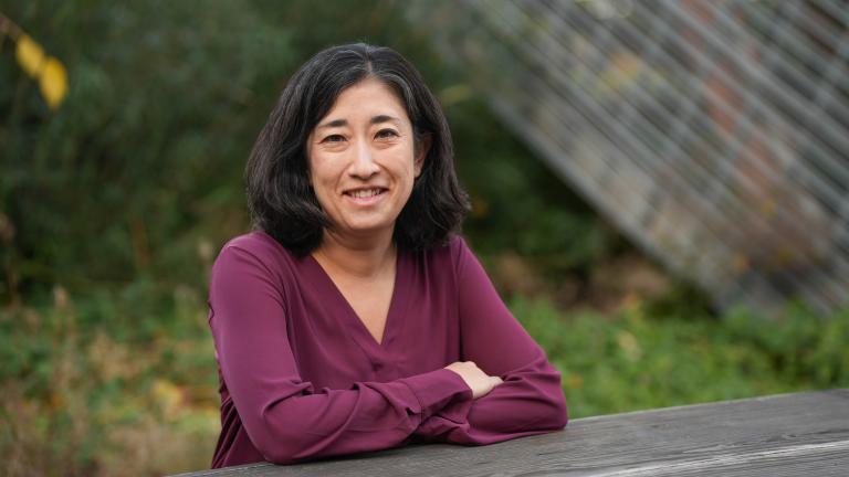June Spector leans forward on a wood surface outside with arms crossed, wearing a maroon blouse. Behind her is a glass and steel sculpture.